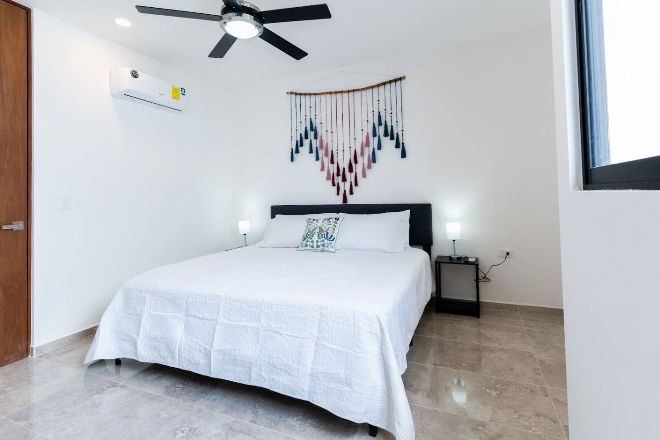 A bedroom with a bed, a ceiling fan, a wall-mounted air conditioner, a bedside table with a lamp, and a decorative wall hanging.