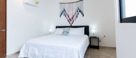 A bedroom with a bed, a ceiling fan, a wall-mounted air conditioner, a bedside table with a lamp, and a decorative wall hanging.