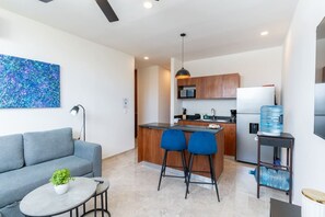 A modern apartment with a grey sofa, a small round table with a plant, a kitchen with wooden cabinets, a built-in island with bar stools, and a large water dispenser. - Atardeceres de Montebello by Kanaa | 1 bd 1.5 bth condo in Northen Merida (Merida)