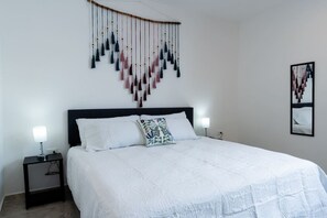 A bedroom with a large bed, two bedside lamps, a wall-mounted hanging decoration, and a framed picture on the wall. - Atardeceres de Montebello by Kanaa | 1 bd 1.5 bth condo in Northen Merida (Merida)