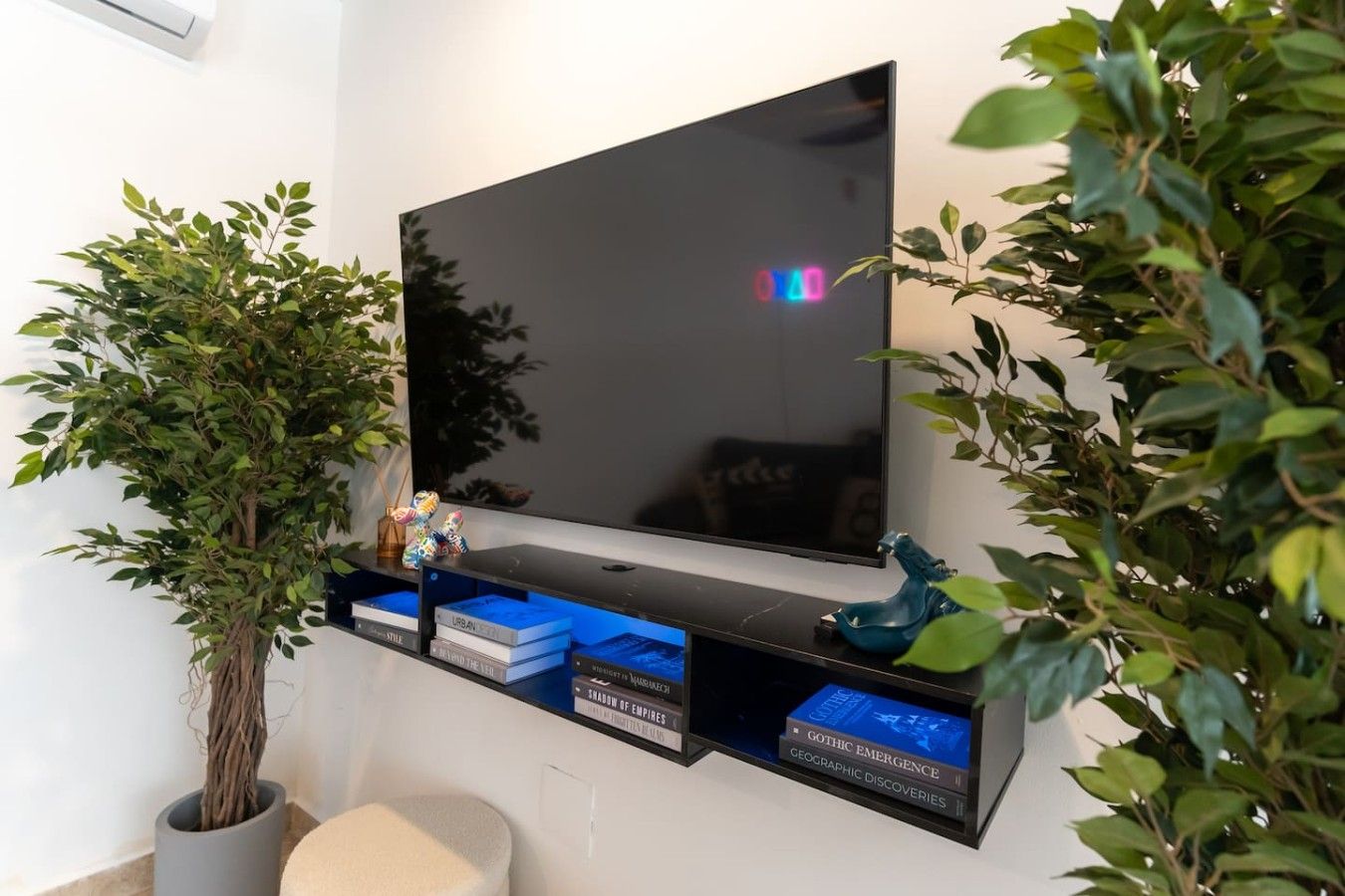A flat-screen TV mounted on a wall with a shelf below it containing books and decorative items.
