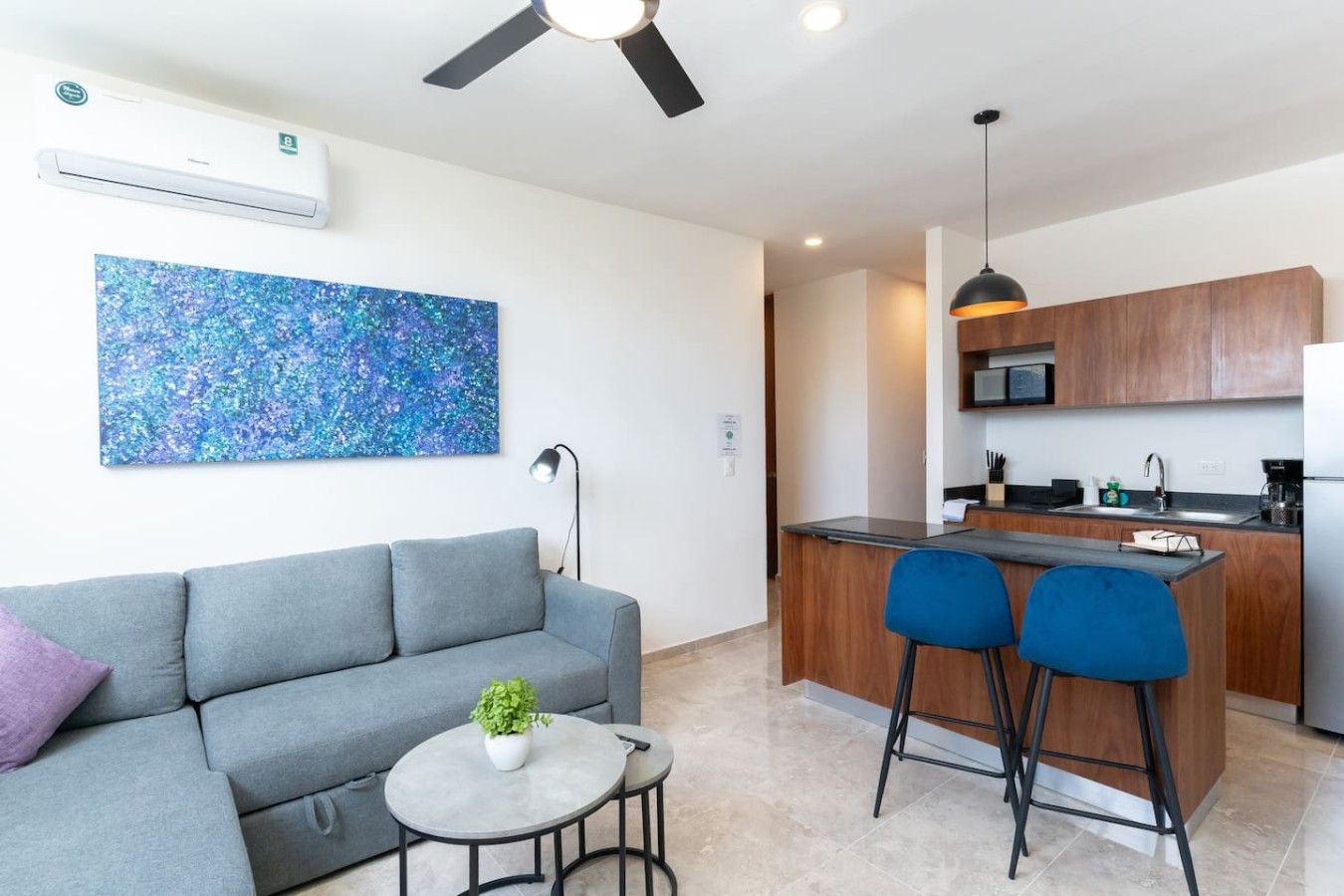 A modern living space with a grey sofa, a small round table with a plant, a blue chair, a kitchen area with wooden cabinets, a microwave, and a ceiling fan.