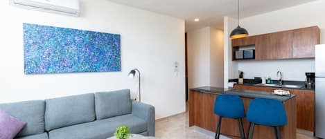 A modern living space with a grey sofa, a small round table with a plant, a blue chair, a kitchen area with wooden cabinets, a microwave, and a ceiling fan.