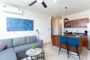 Um espaço de estar moderno com sofá cinzento, uma pequena mesa redonda com uma planta, uma cadeira azul, área de cozinha com armários de madeira, micro-ondas e ventoinha de teto.
