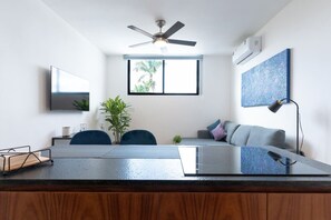 A modern living room with a dining area, a ceiling fan, a large window, and a mounted artwork. - Atardeceres de Montebello by Kanaa | 1 bd 1.5 bth condo in Northen Merida (Merida)