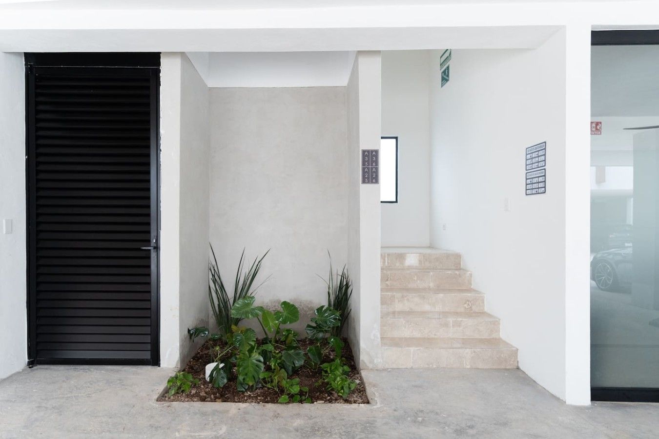 Een modern gebouw met een entree, een zwarte deur, een kleine tuin met planten, en een trap die naar een hoger gelegen niveau leidt.