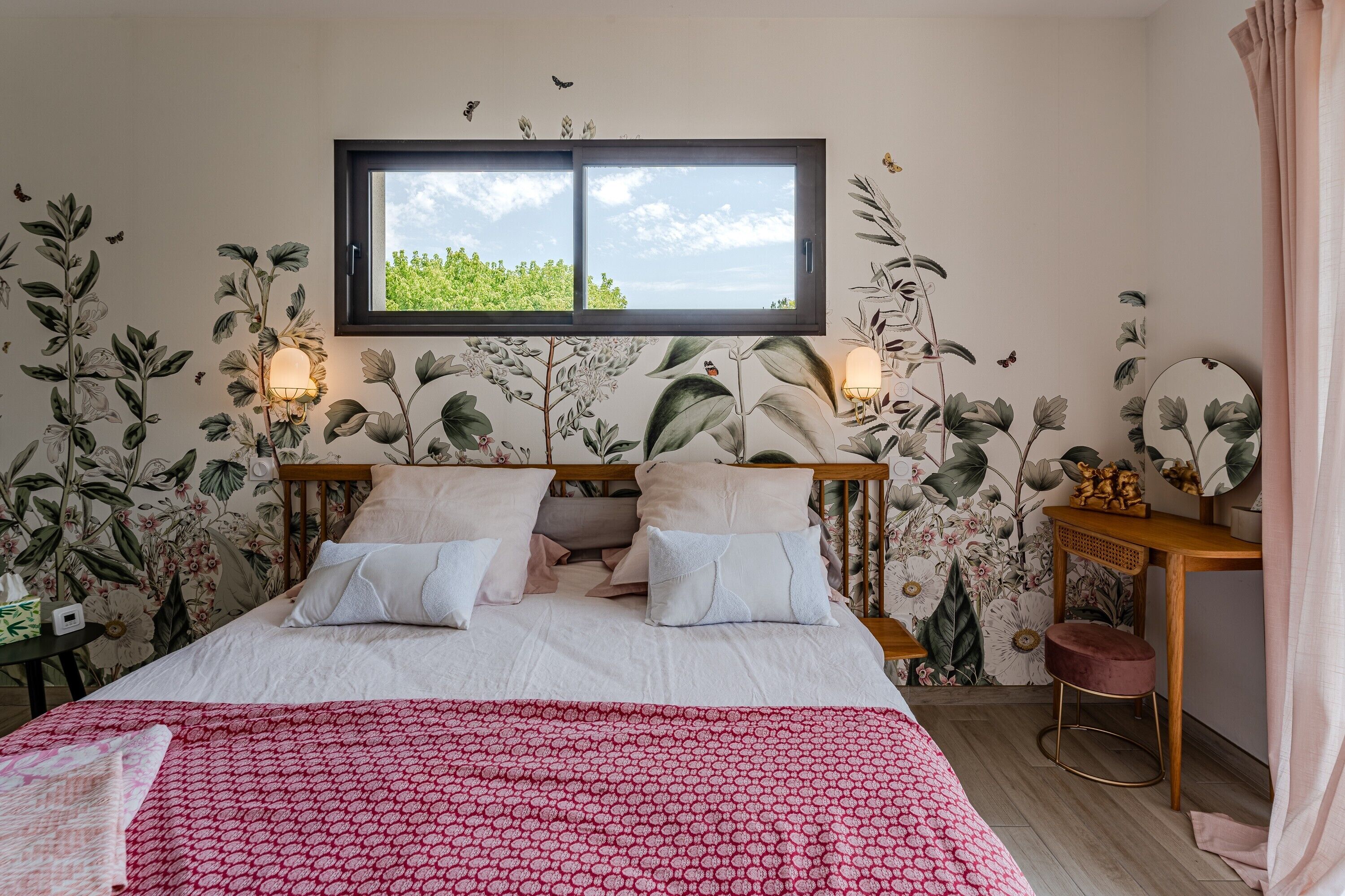 Une chambre à coucher avec un lit, une fenêtre, une table de chevet et un mur recouvert d’un papier peint à motifs floraux.