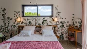 Une chambre à coucher avec un lit, une fenêtre, une table de chevet et un mur recouvert d’un papier peint à motifs floraux.