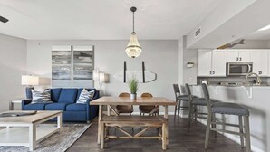 A modern living room with a blue sofa, a wooden dining table, and a kitchen area with white cabinets and a microwave. - Cozy Retreat in the Sky (Destin)