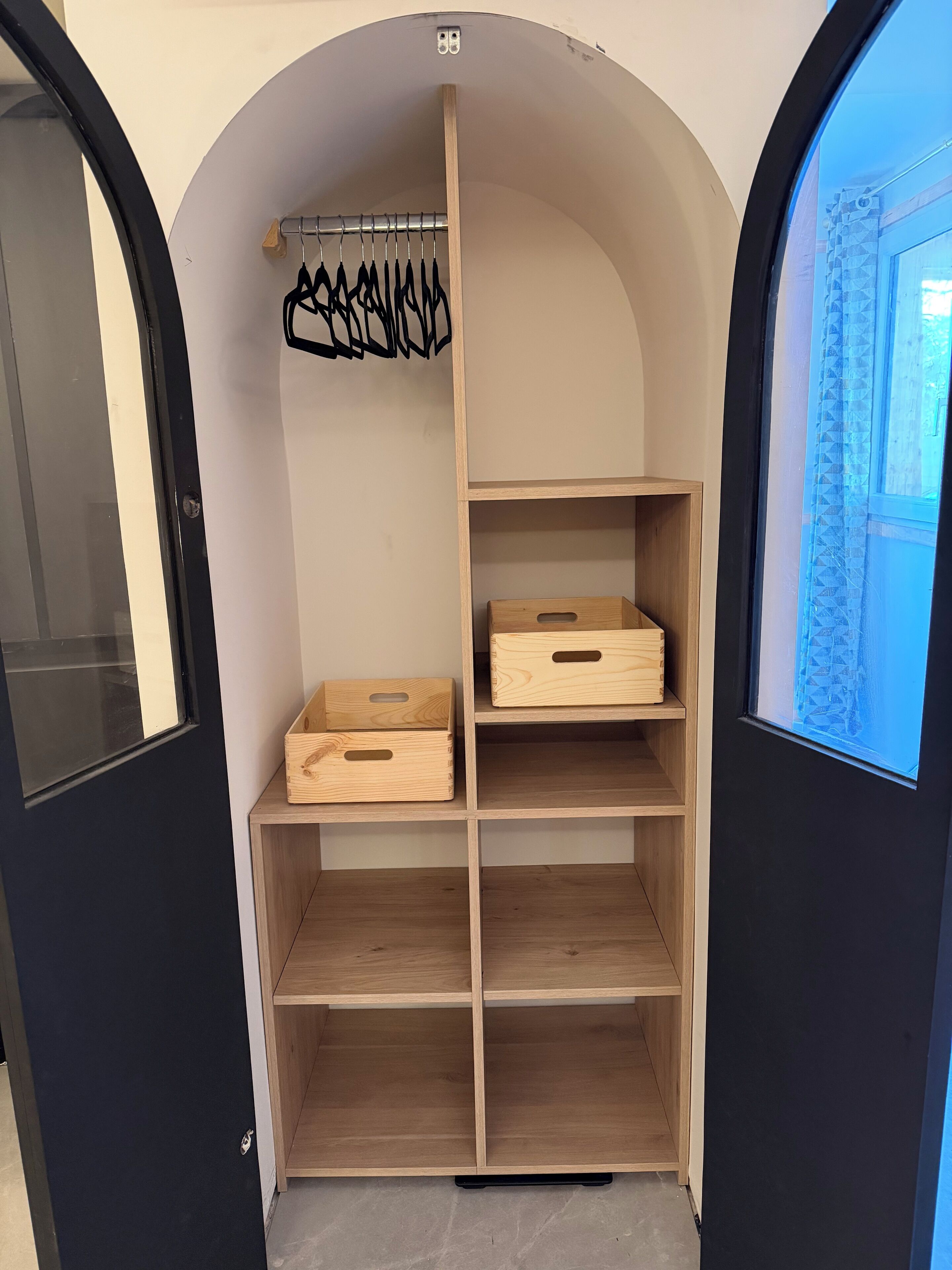 Ein kompakter, bogenförmiger Holzschrank mit mehreren Ablagen und Kleiderstange, vermutlich zur Aufbewahrung von Gepäck oder persönlichen Gegenständen.