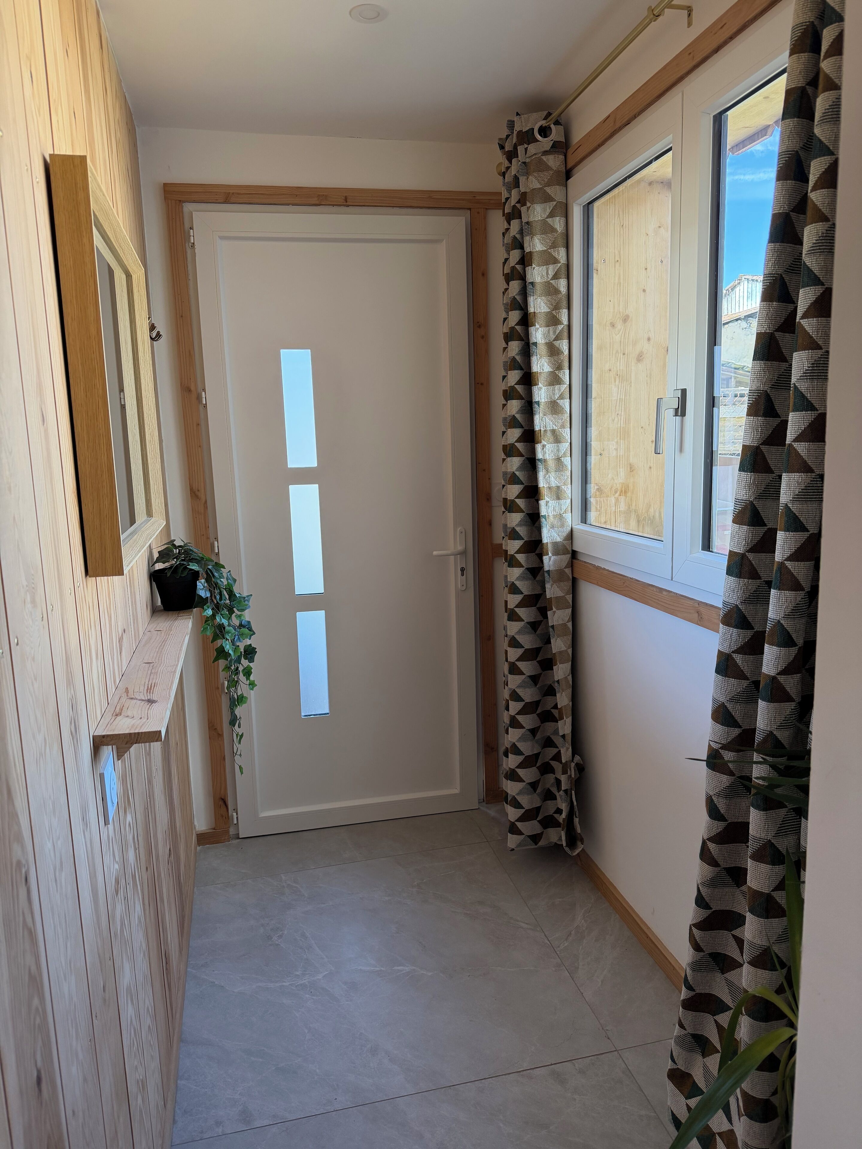 A narrow hallway with a wooden door, a window with patterned curtains, and a potted plant.