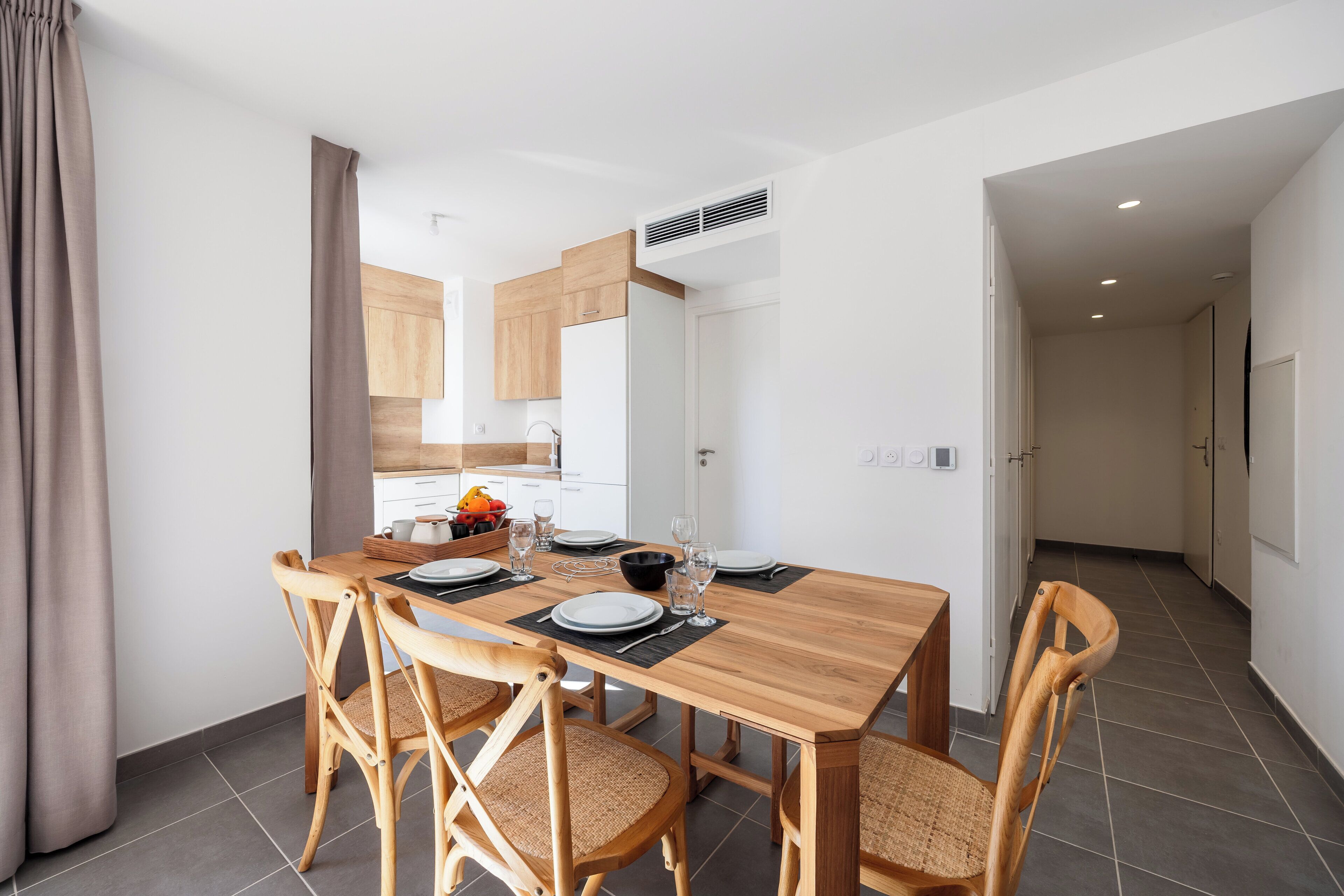 A dining area with a wooden table set for four, surrounded by wicker chairs. A kitchen is visible in the background.