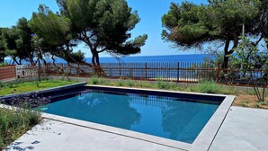 A rectangular swimming pool with a tiled deck, surrounded by a wooden fence, with a view of the sea and trees in the background. - La Corniche Bleue (Sausset-les-Pins)