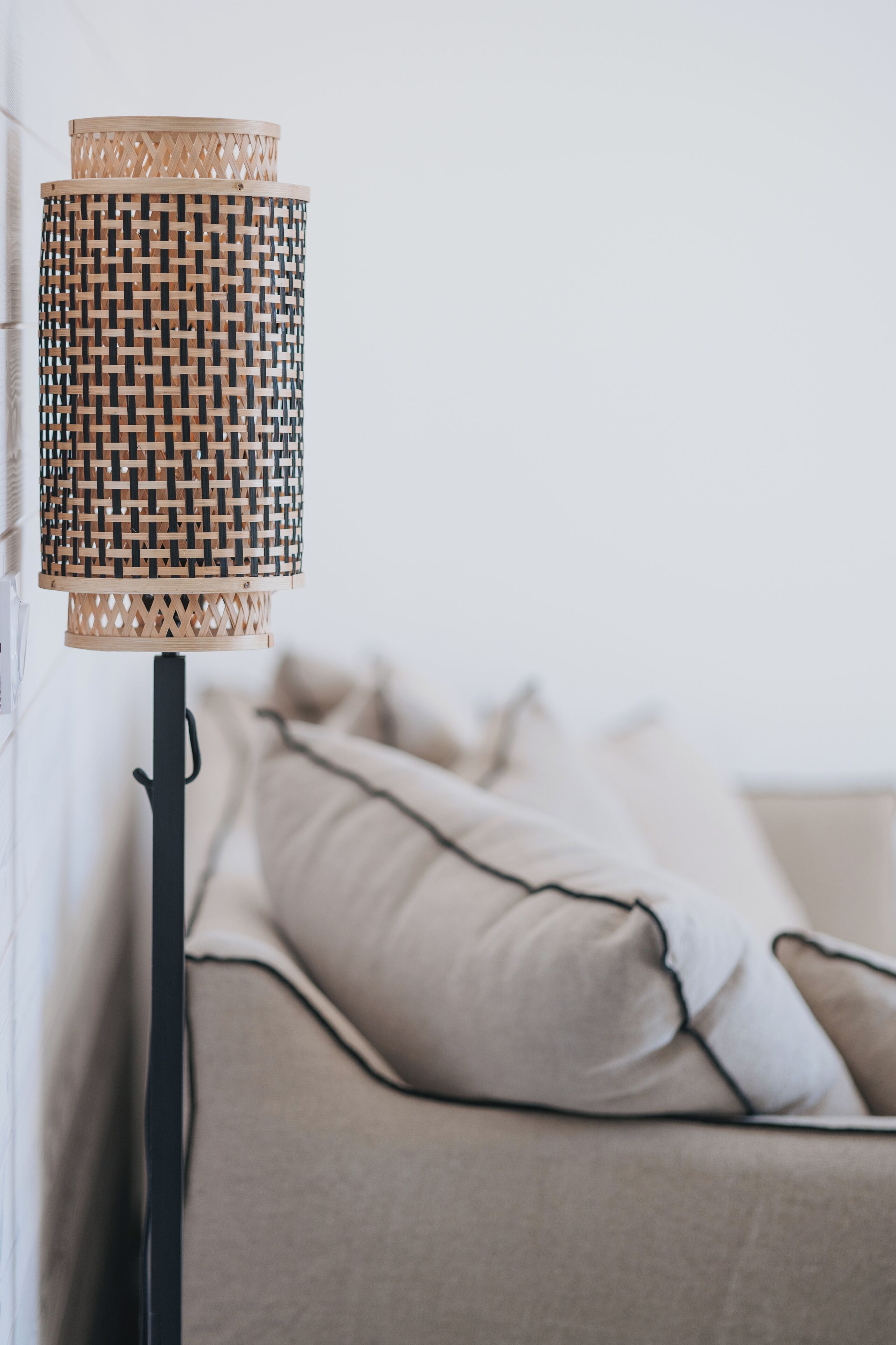 Une lampe avec un abat-jour en osier, un pied noir et un canapé beige agrémenté de coussins, le tout dans une pièce.