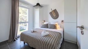 A modern bedroom with a large bed, a hanging light fixture, and a view of the outdoors. - La Corniche Bleue (Sausset-les-Pins)