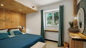Een slaapkamer met een houten hoofdeinde, een bed met blauw beddengoed, een bankje, een raam met gordijnen en een rustieke houten dressoir met spiegel.