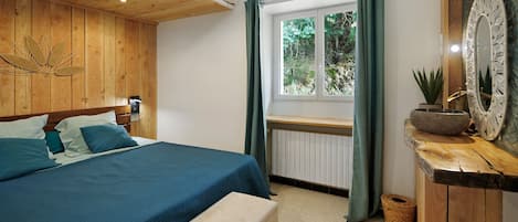 Un dormitorio con cabecera de madera, cama con ropa de cama azul, una banca, una ventana con cortinas y un aparador rústico de madera con espejo.