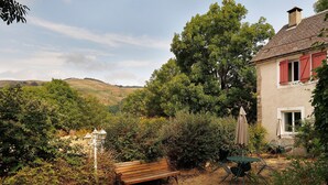 Ein rustikaler Sitzbereich im Freien mit einer Bank und einem Tisch, umgeben von üppiger Vegetation und einer sanften Hügellandschaft im Hintergrund.