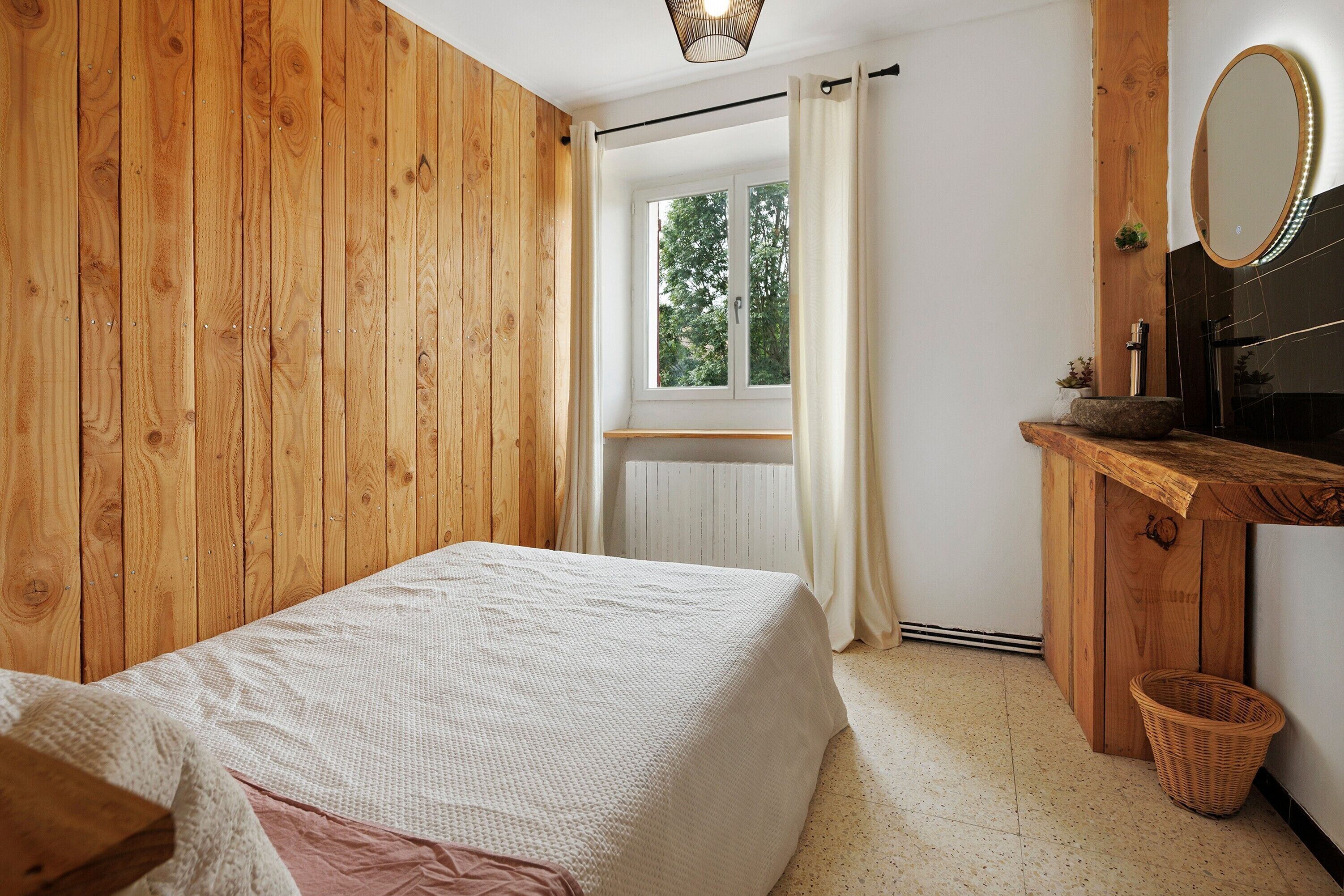 A bedroom with a wooden wall, a bed, a window with curtains, a mirror, and a small wooden dresser.