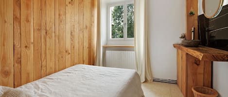A bedroom with a wooden wall, a bed, a window with curtains, a mirror, and a small wooden dresser.