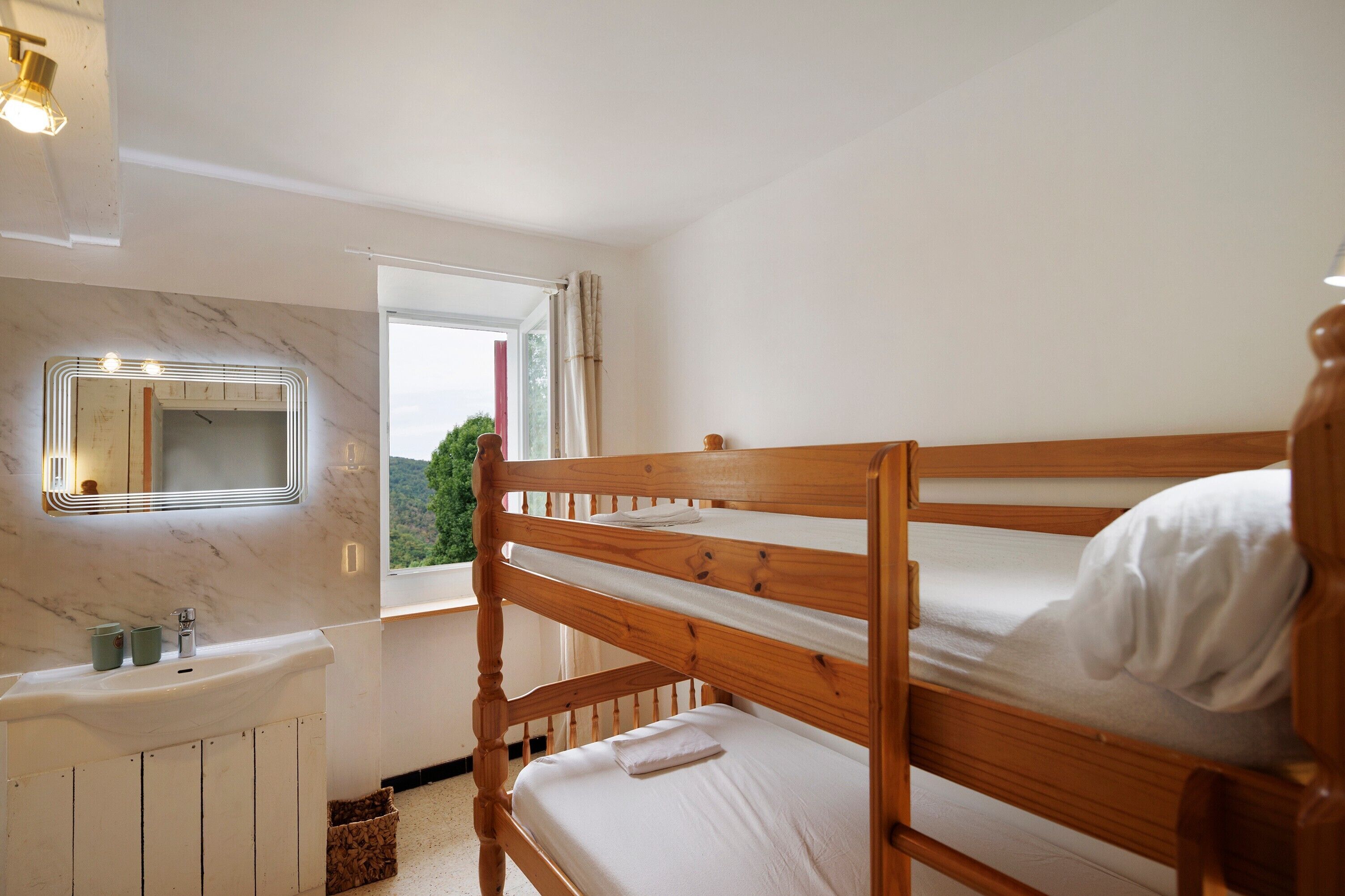 Una cama litera con estructura de madera y ropa de cama blanca, una mesita de noche con espejo y una ventana que da a vistas de vegetación.