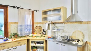 A compact kitchen with a gas stove, sink, and wooden cabinets. There is a window with a curtain, and a shelf with dishes and bottles. - Residence with swimming pool in Santa Margherita di Pula (Santa Margherita di Pula)