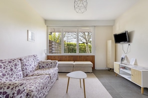 A modern living room with a sofa, a small table, a TV, and a window with a view of greenery. - Mirabel Cottage at Château Saint Ahon (Blanquefort)