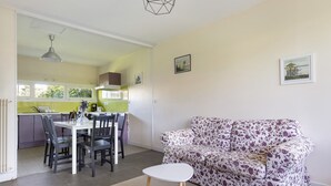 A living room with a sofa, a dining table with chairs, and a kitchen in the background. - Mirabel Cottage at Château Saint Ahon (Blanquefort)