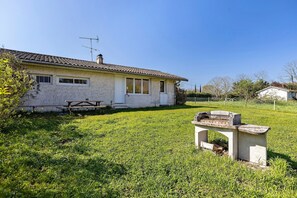 Une maison avec un toit en tuiles, une cheminée et une cour verdoyante équipée d’un barbecue.