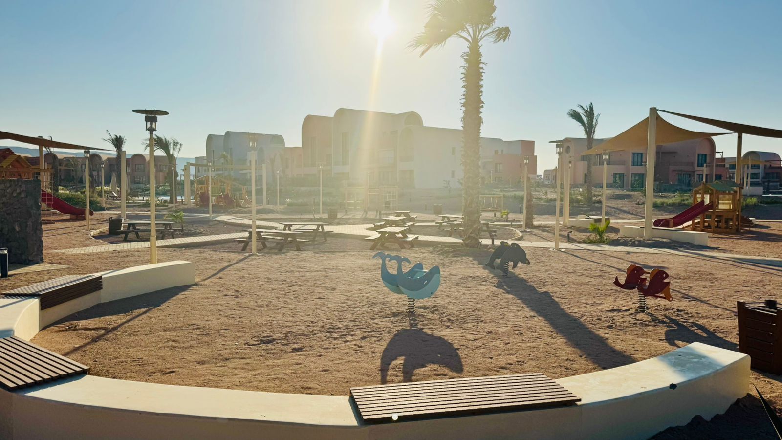 A playground with swings and slides, surrounded by buildings and palm trees.