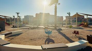 A playground with swings and slides, surrounded by buildings and palm trees.