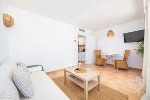 Una sala de estar moderna con un sofá, una mesa de centro, sillas de mimbre, un televisor y una cocina al fondo.