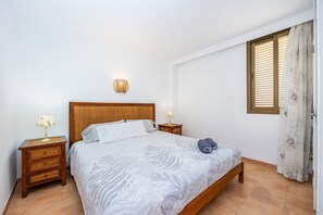Une chambre à coucher avec un lit, une table de chevet avec une lampe, un luminaire fixé au mur et une fenêtre avec des stores.