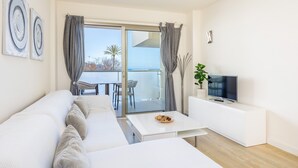 A modern living room with a sofa, a coffee table, a TV on a white cabinet, and a balcony with a view. - Incredible Sea Views – 50 m from Cala Millor Beach (Cala Millor)