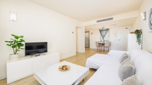 A modern living room with a flat-screen TV, a white sofa, a coffee table, and a dining area in the background. - Incredible Sea Views – 50 m from Cala Millor Beach (Cala Millor)