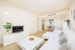 Un salon moderne avec un téléviseur à écran plat, un canapé blanc, une table basse et un coin repas à l’arrière-plan.