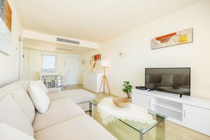 A modern living room with a sofa, TV, and a dining area in the background. - Seafront Apartment in Cala Millor – Mallorca (Cala Millor)