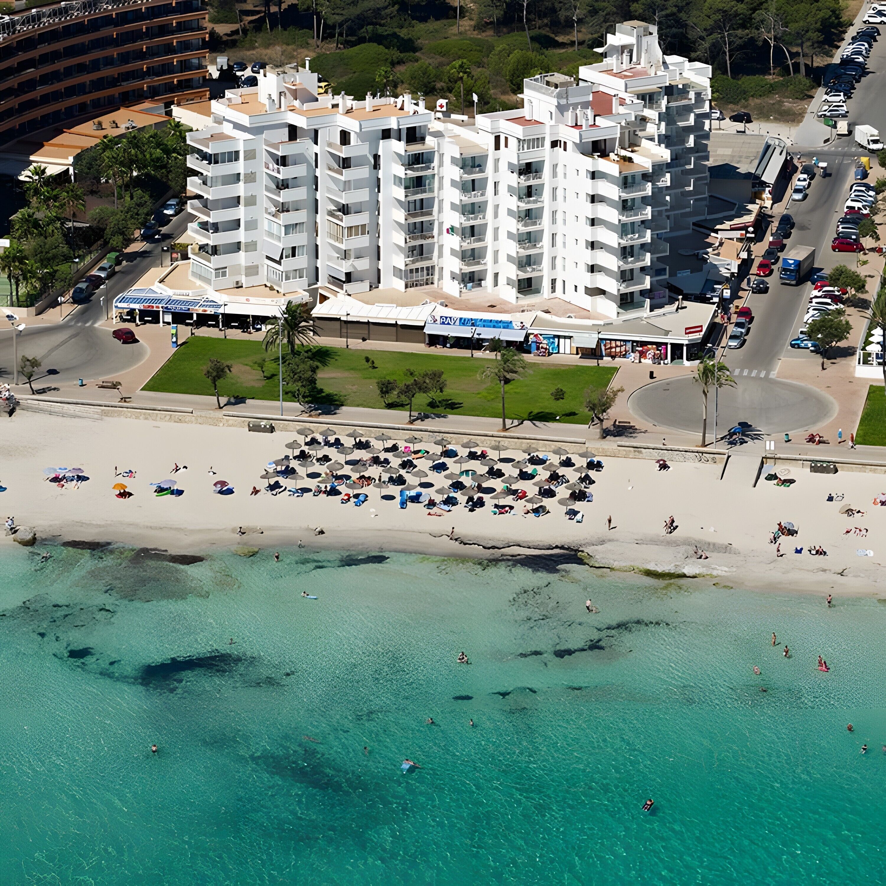 Ein Luftbild eines Küstenhotels mit mehreren Stockwerken, einem Strand mit Sonnenschirmen und klarem, türkisblauem Wasser.