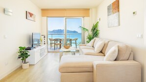 Un soggiorno moderno con un divano, una televisione, un tavolo da pranzo con sedie, una pianta in vaso e un balcone con vista sul mare.