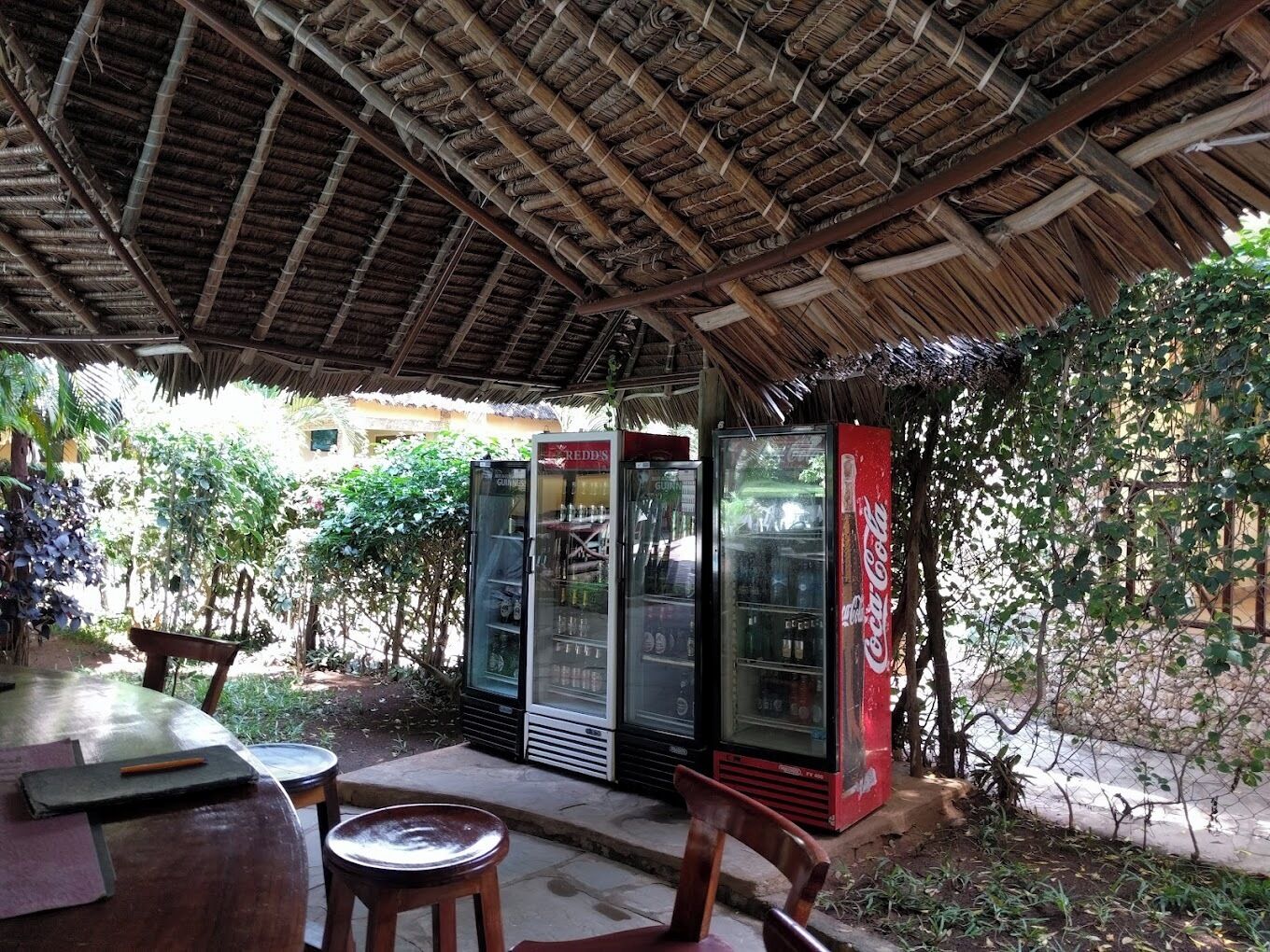 Un área de estar al aire libre con techo de paja, una mesa y dos sillas. Hay dos refrigeradores, uno con el logotipo rojo de Coca-Cola y otro con el logotipo de Guinness.