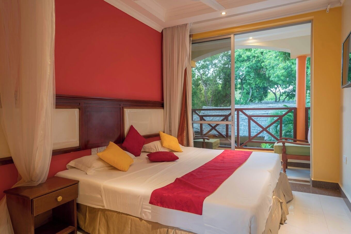 A hotel room with a large bed, a nightstand, and a view of a balcony with outdoor furniture.