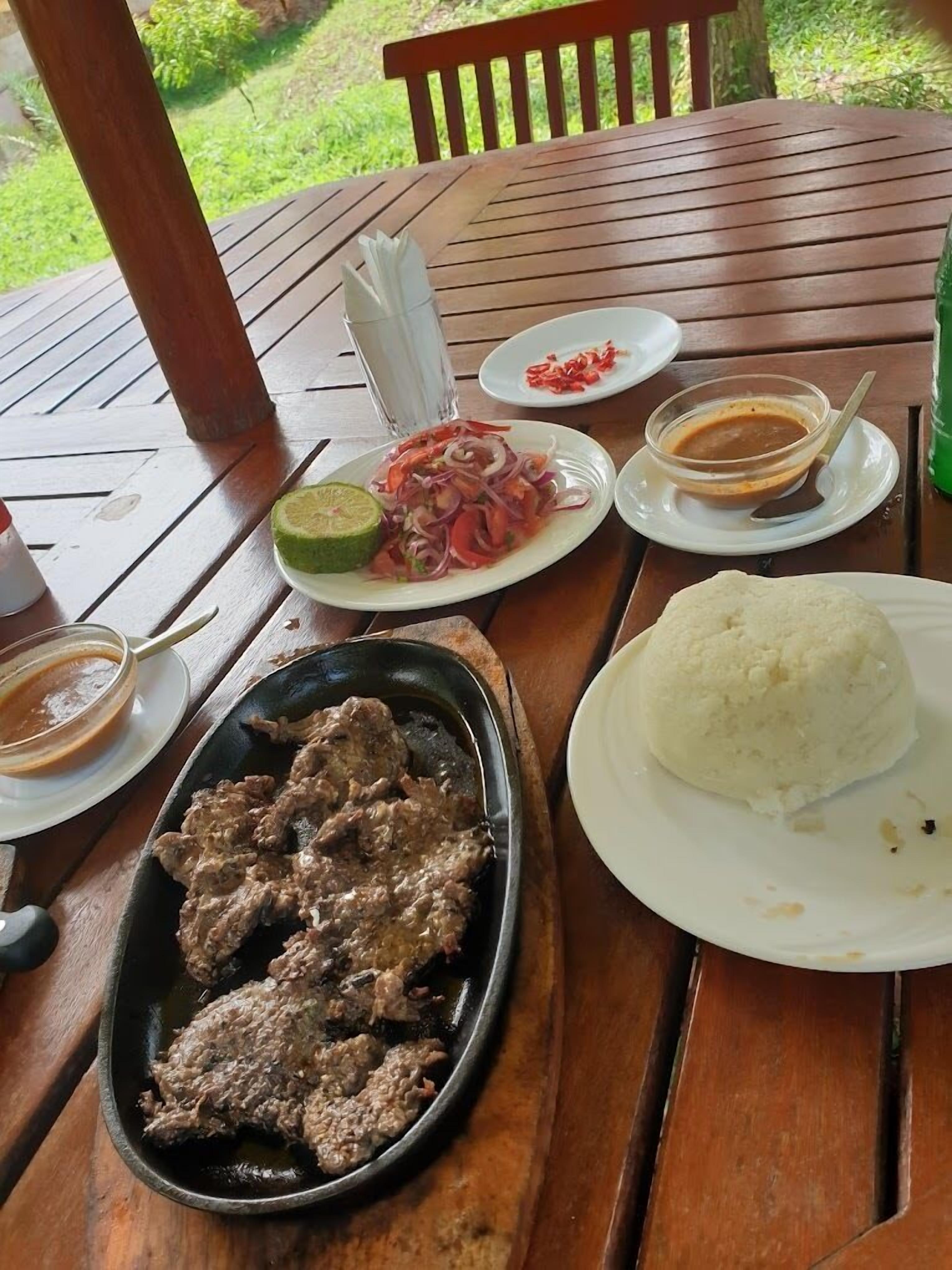 A wooden table with a meal consisting of grilled meat, rice, a salad with onions and peppers, and a small bowl of sauce.