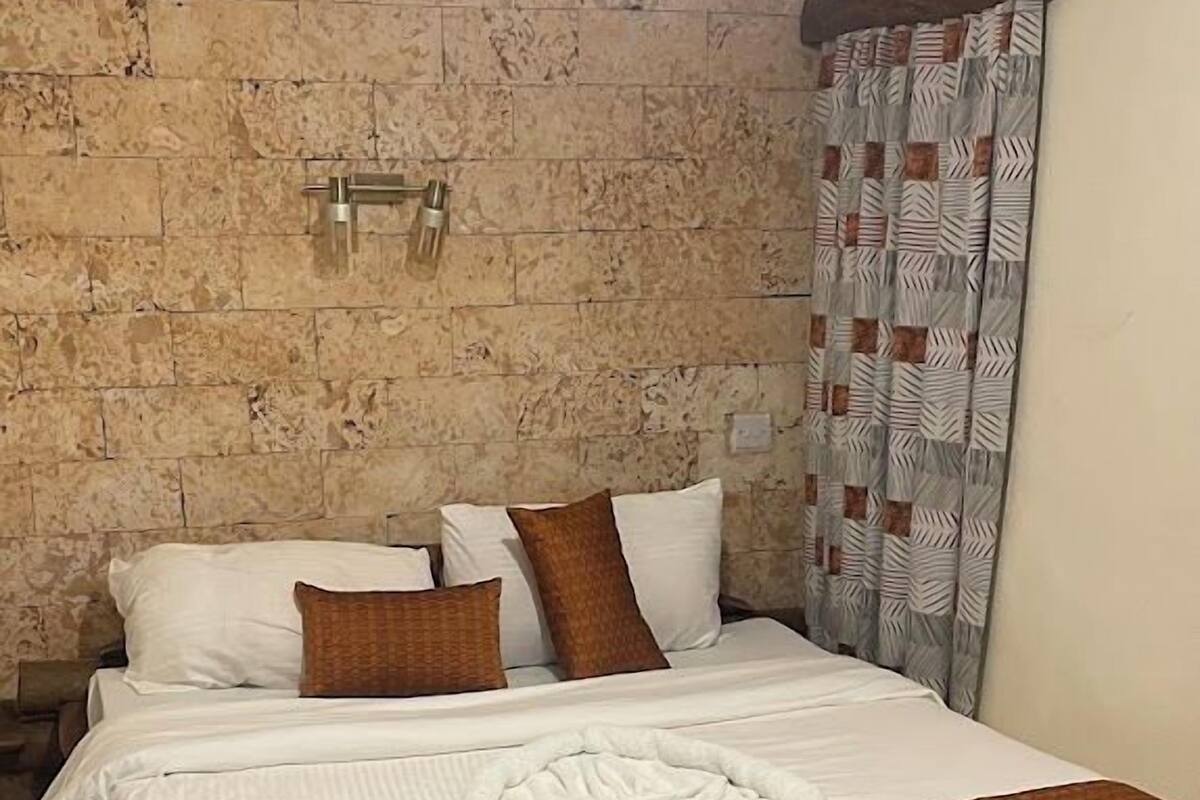 A neatly made bed with white linens and brown accents, a stone wall, and a patterned curtain.