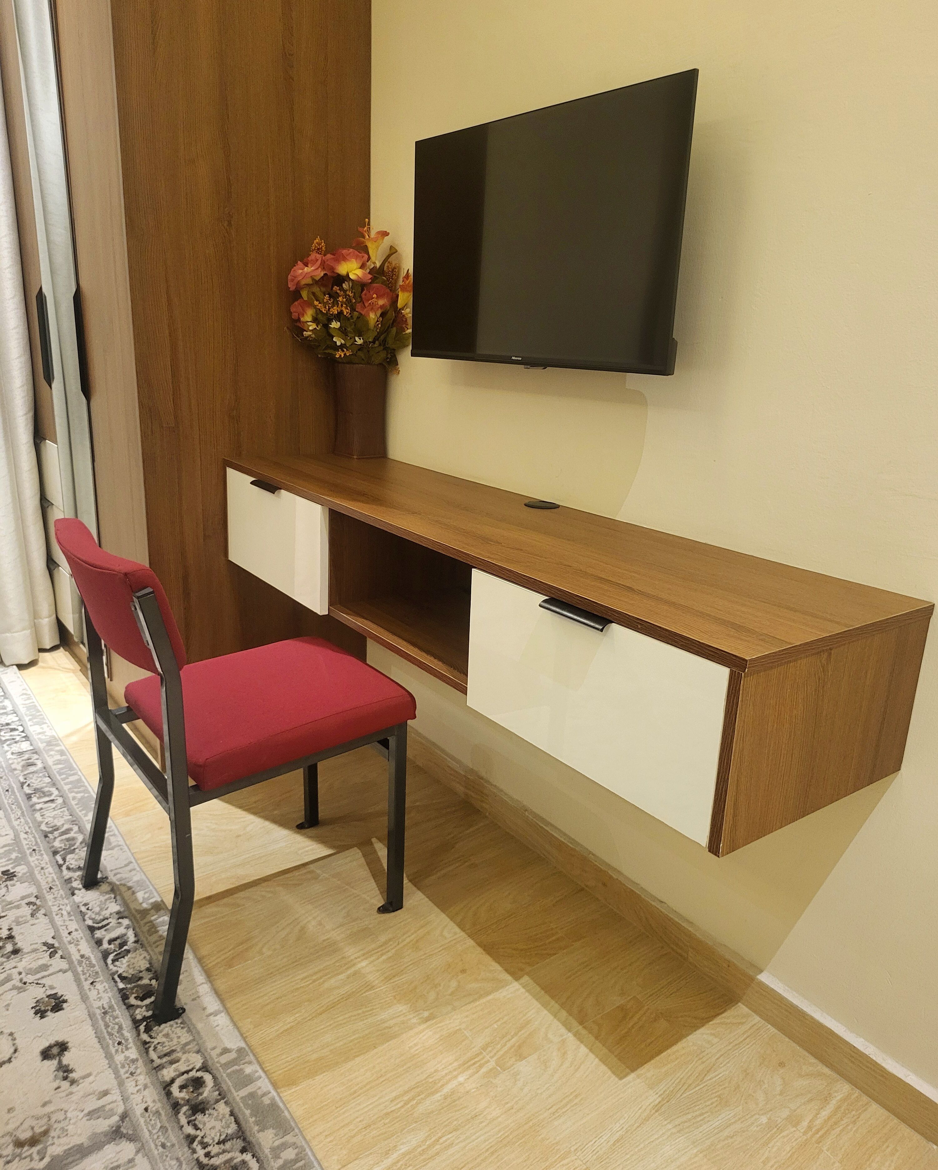 Een kamer met een houten bureau, een rode stoel, een aan de muur bevestigde televisie en een plant in een pot.