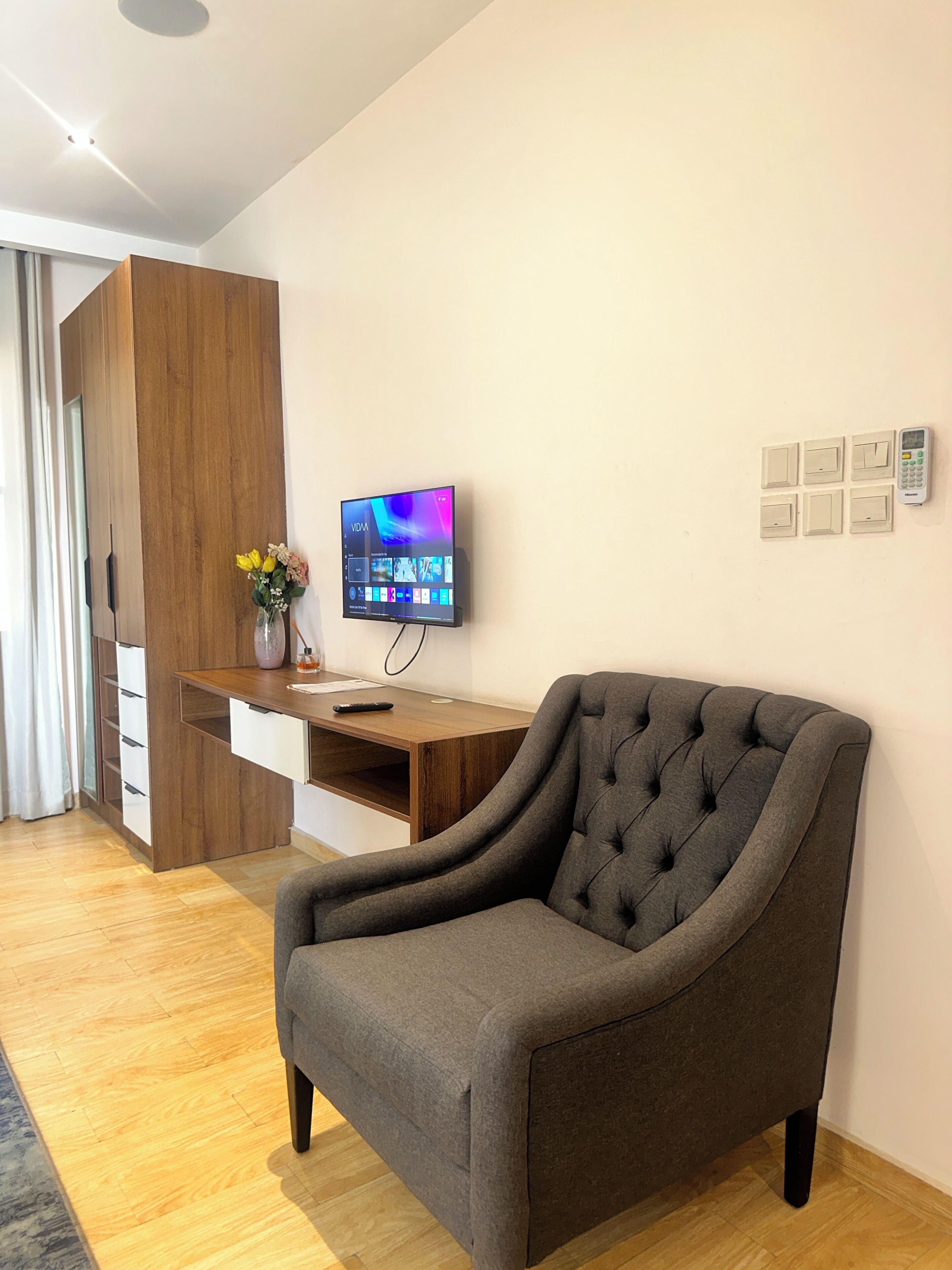 A modern room with a grey armchair, a wooden desk with a flat-screen TV, and a wall-mounted control panel.