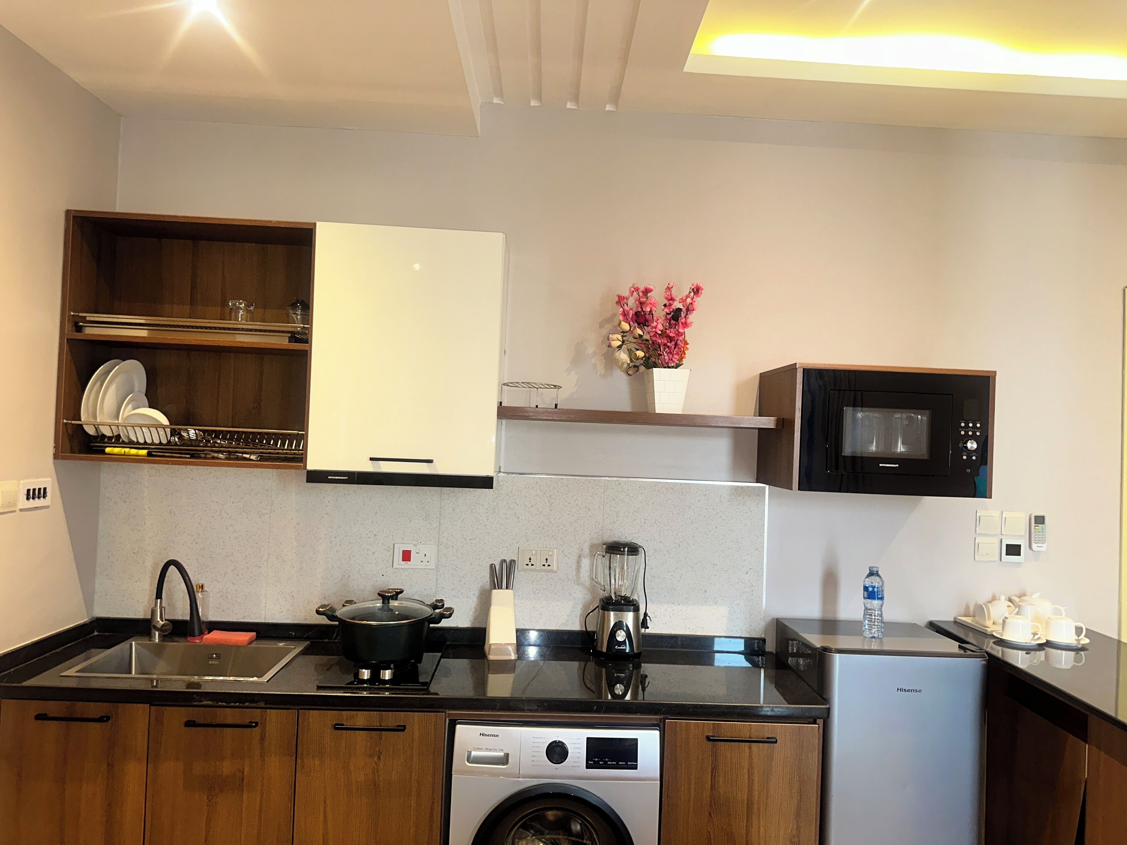 A compact kitchen with wooden cabinets, a microwave, a sink, a stove, a blender, and a washing machine.
