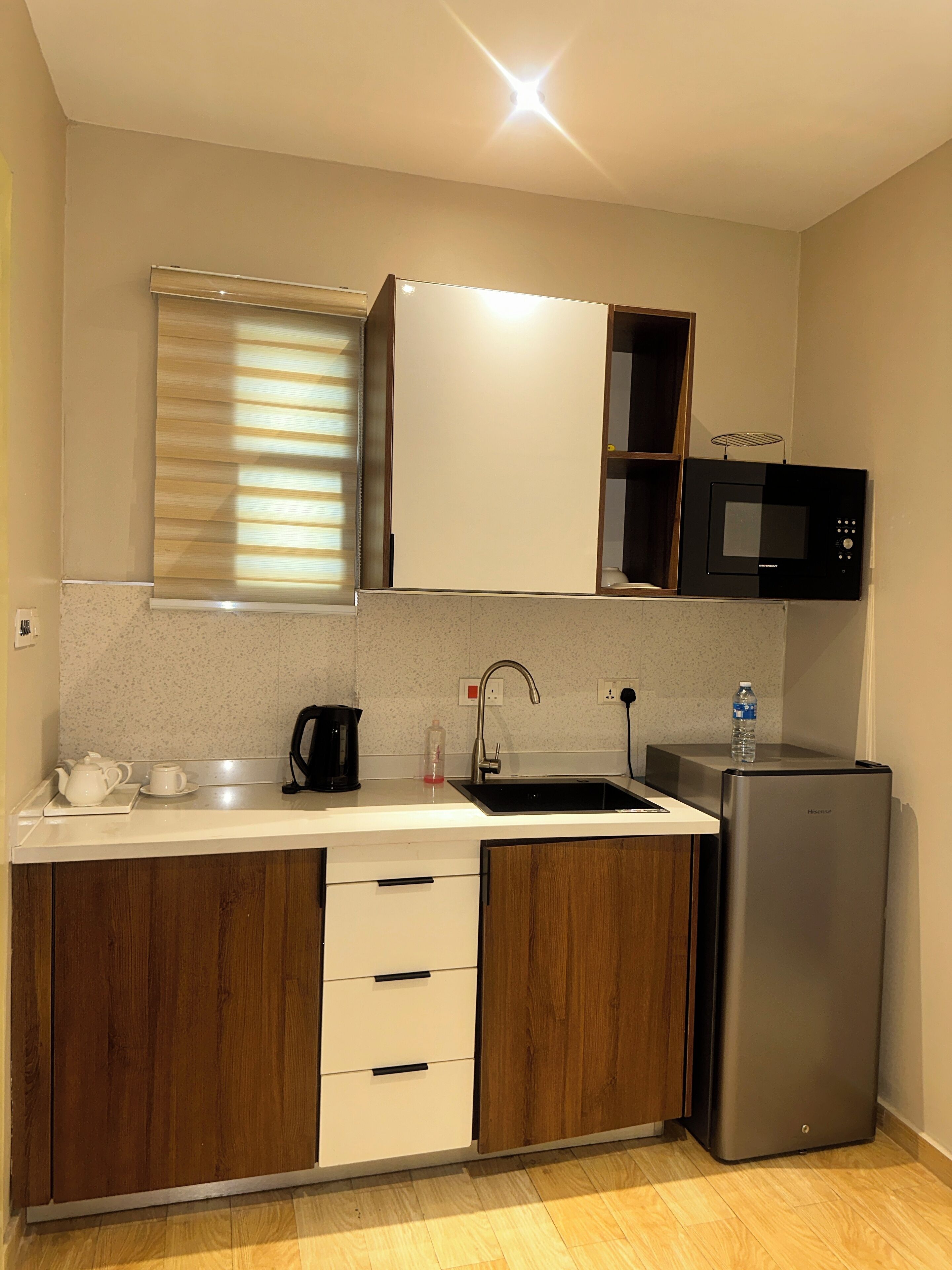 A compact kitchen with a sink, a microwave, a refrigerator, wooden cabinets, and a window with blinds.