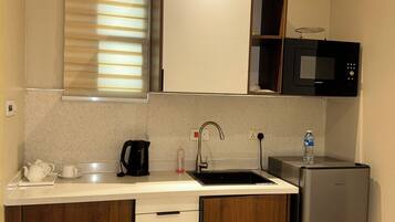 A compact kitchen with a sink, a microwave, a refrigerator, wooden cabinets, and a window with blinds.