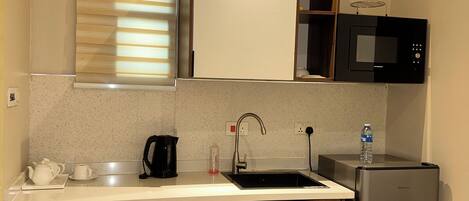 Cocina compacta con fregadero, microondas, refrigerador, gabinetes de madera y una ventana con persianas.