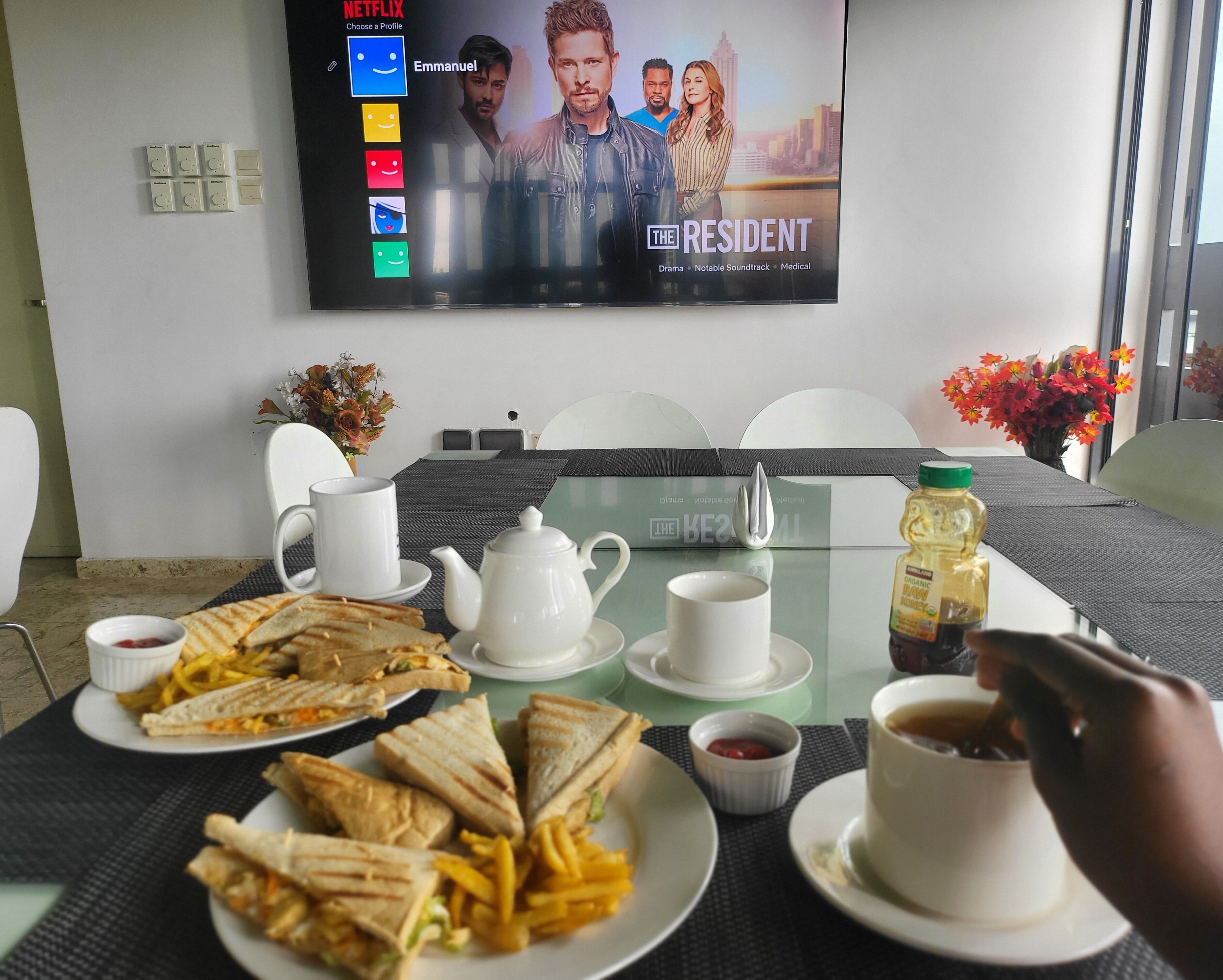 A hotel room with a bed, a TV displaying a show, a dining table with food and drinks, and a vase with flowers.